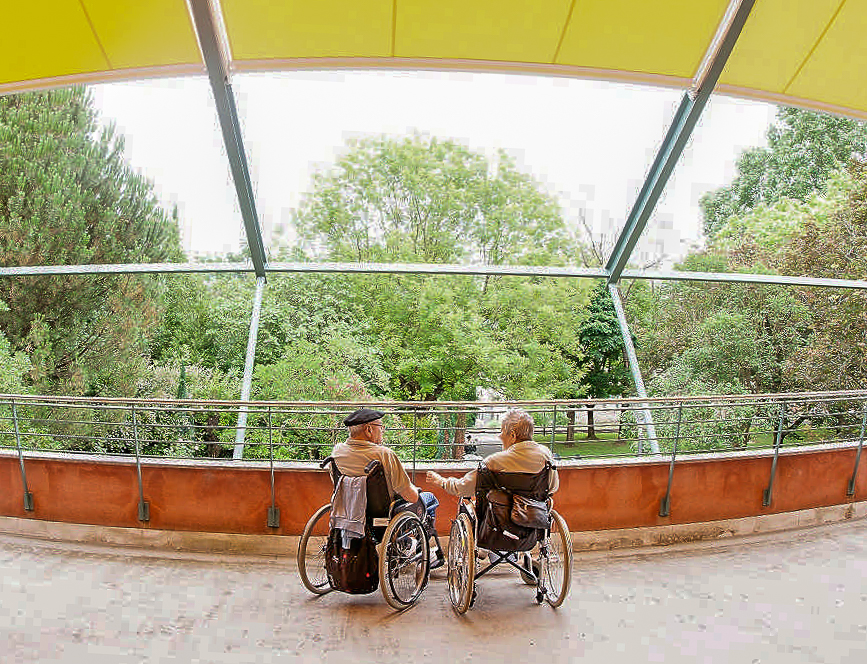 una pareja de residentes sentados en sus sillas de ruedas contemplan el jardín desde la terraza