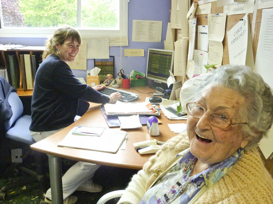 una enfermera trabaja frente al ordenador mientras atiende a una residente que se ve en primer plano, ambas sonríen cómplices